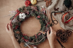 Rustic Christmas wreath flat lay. Hands holding fir branches, red berries and pine cones,thread, scissors, cinnamon, cotton on rustic wooden background. Making  wreath at workshop