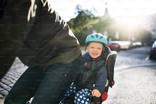 A small toddler boy with helmet sitting in bicycle seat with unrecognizable father outdoors on a street in city.