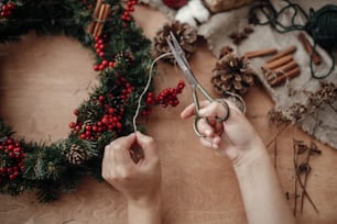 Hands making rustic christmas wreath, cutting rope with  scissors at fir branches, red berries,pine cones on rustic wooden background. Atmospheric moody image at holiday workshop