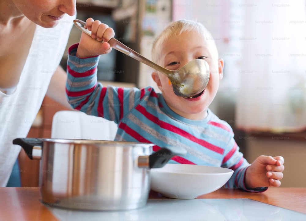 幸せな障害のあるダウン症の男の子が、室内でお玉からスープを食べ、ランチタイム。