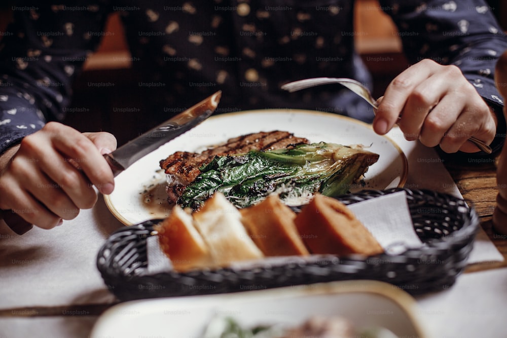 Hands holding fork and knife and eating delicious juicy steak with grilled cabbage,tomatoes and cheese on table at cafe in city street. Man tasting bbq with vegetables in restaurant