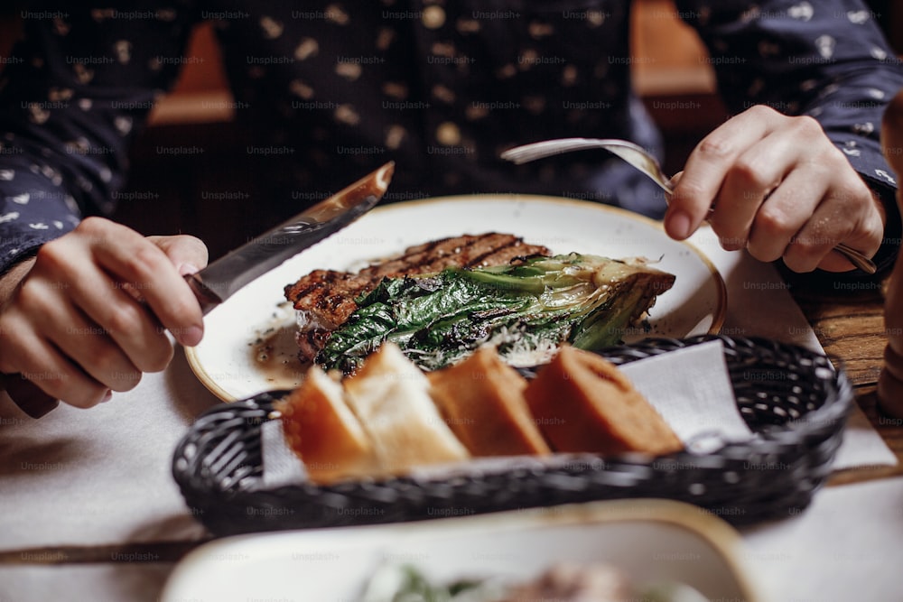 Les mains tenant une fourchette et un couteau et mangeant un délicieux steak juteux avec du chou grillé, des tomates et du fromage sur la table d’un café dans la rue de la ville. Homme dégustant un barbecue avec des légumes dans un restaurant