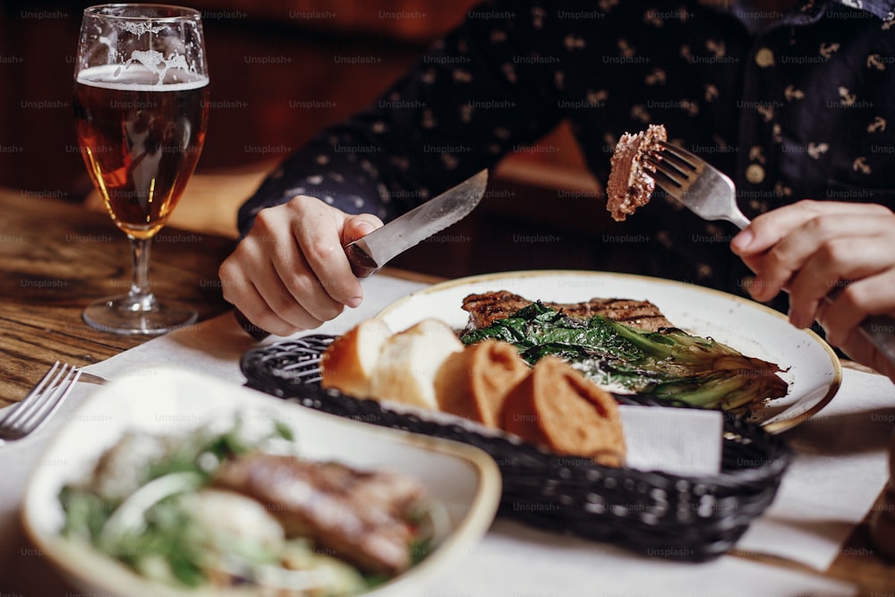 Homme mangeant un délicieux steak juteux avec du chou grillé, des tomates et du fromage sur la table à la terrasse du café dans la rue de la ville. Mains tenant une fourchette et un couteau et dégustant un barbecue avec des légumes dans un restaurant
