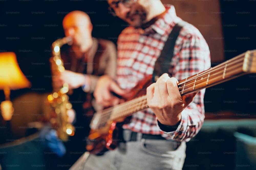 Close up of talented bass guitarist playing his instrument. In background saxophonist playing sax. Home studio interior.