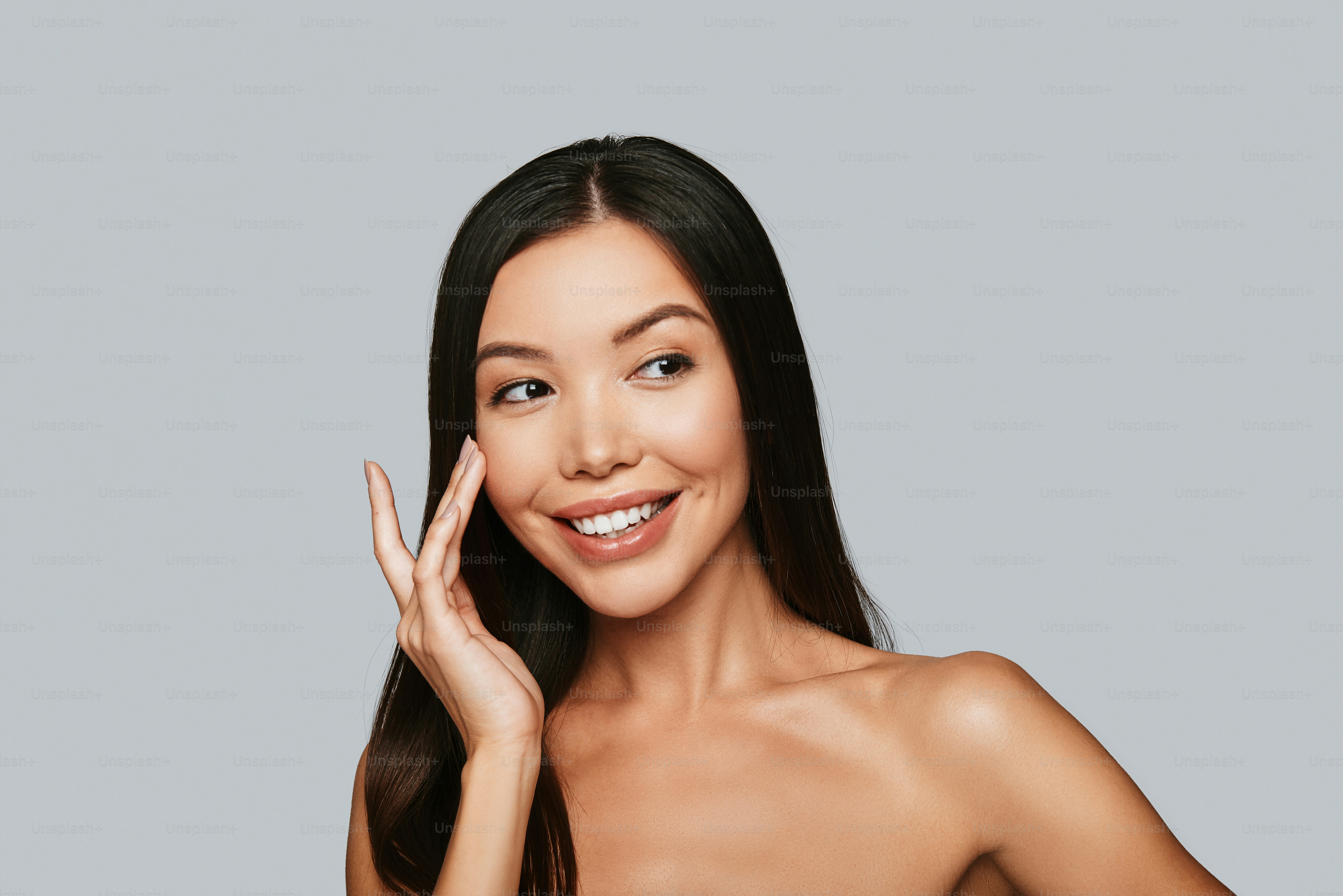 Beautiful young Asian woman looking away and smiling while standing against grey background