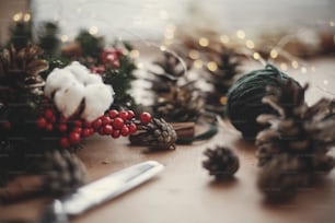 Fir branches, wreath, red berries, pine cones, thread, scissors, cinnamon, cotton, lights on rustic wooden background. Details for making christmas wreath at workshop. Atmospheric image