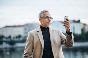 Mature handsome businessman with smartphone standing by river Vltava in Prague city, taking selfie.