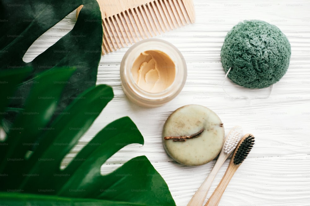Eco friendly natural bamboo toothbrushes, shampoo bar, toothpaste in glass, wooden brush and konjaku sponge on white wood with green monstera leaves. Zero waste flat lay