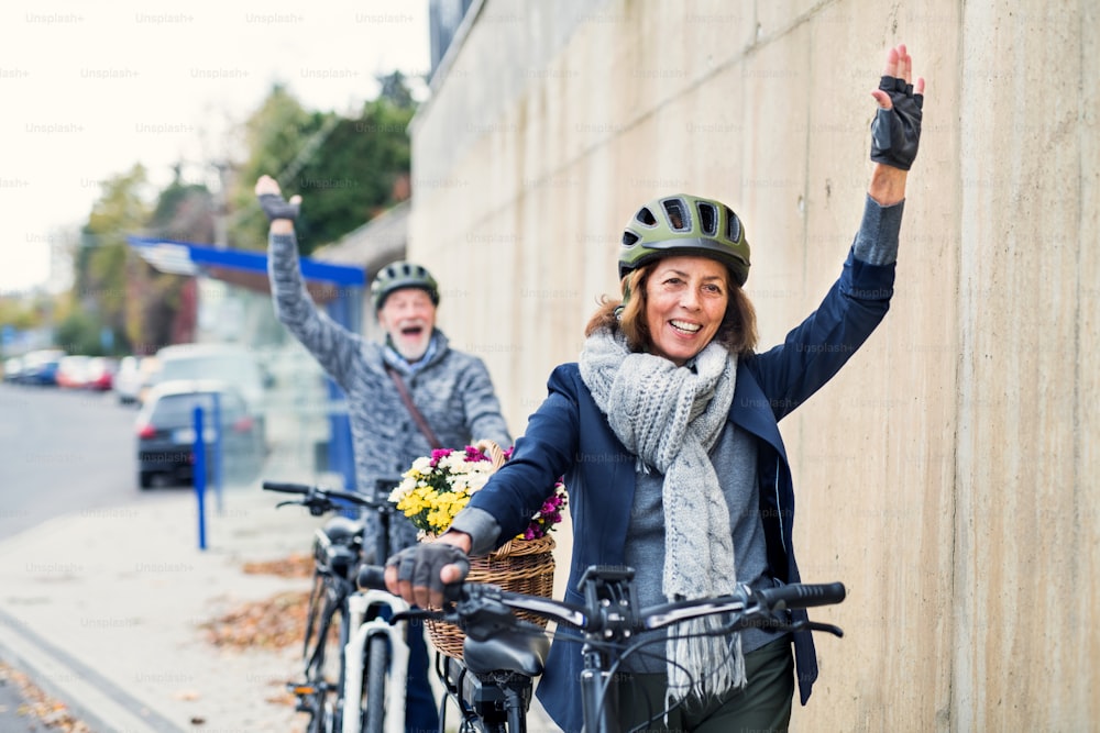 Couple de personnes âgées actives avec des casques et des vélos électriques debout à l’extérieur sur une route de la ville, saluant quelqu’un.