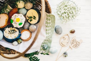 Easter basket with stylish eggs, easter bread, ham, sausage, butter, and flowers, easter wooden bunny and chicken decorations on rustic wood, top view. Happy Easter concept