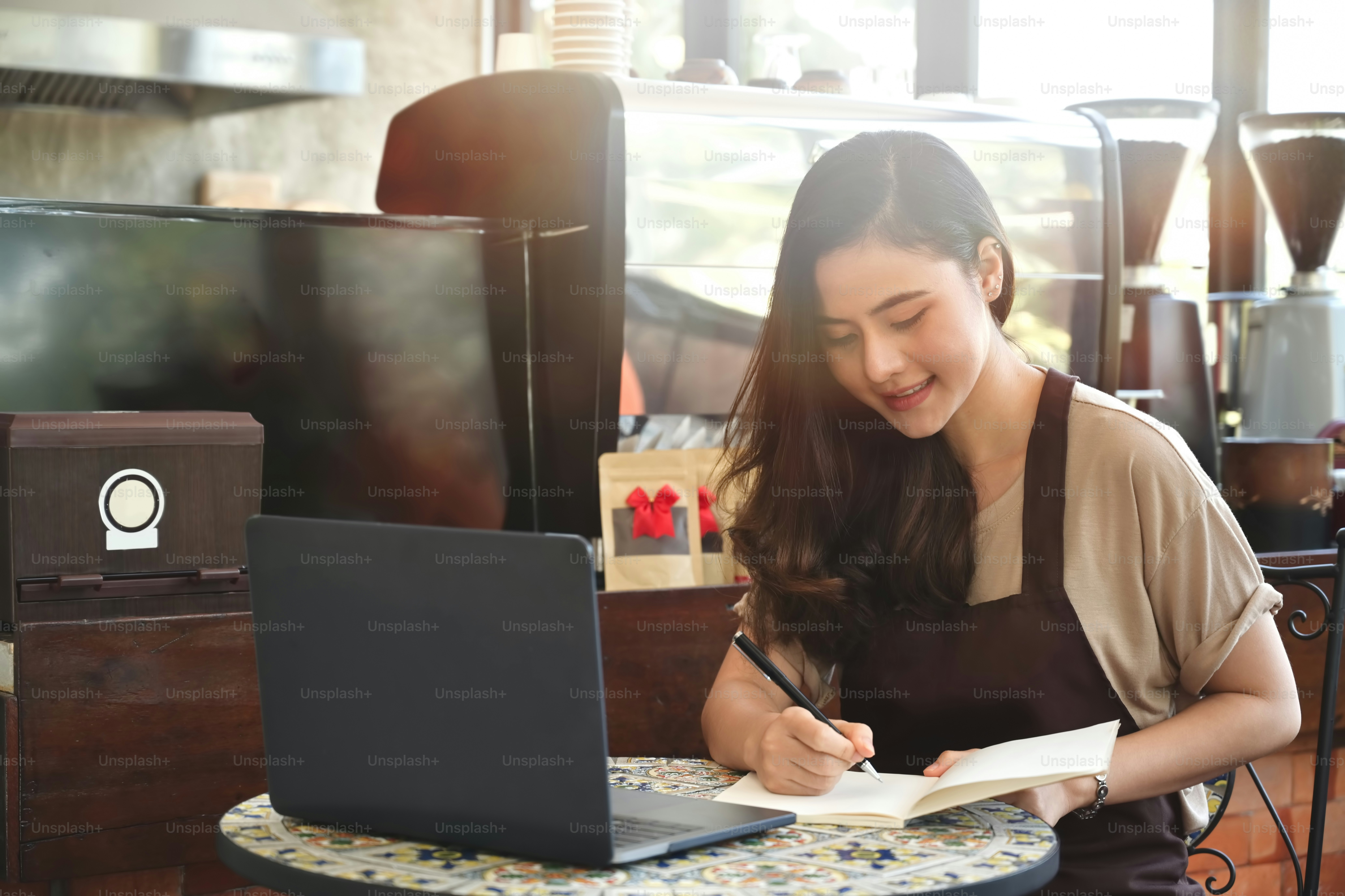 Young Asian barista in apron and white shirt writing in notebook at coffee shop.