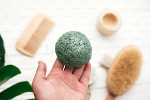 Hand holding natural konjaku sponge on background of bamboo brush, deodorant in glass on white wood with green monstera leaves. Zero waste concept. Choose plastic free eco products