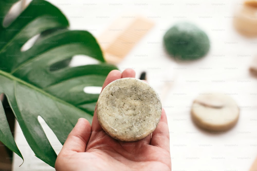 Zero waste. Choice plastic free eco products. Hand holding natural solid shampoo bar on background of bamboo brush, deodorant, sponge on white wood with green monstera leaves.