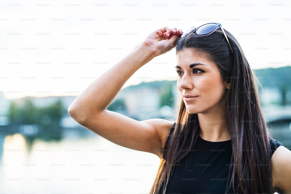 A beautiful woman in black dress standing by a river in city of Prague, holding sunglasses.