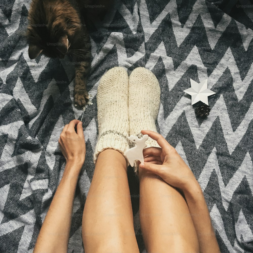 クリスマスウールの靴下とスタイリッシュなトナカイのおもちゃを手にした女の子の足で休日の飾りで遊ぶかわいい猫。上面図。雰囲気のある居心地の良いイメージ、暖かい冬の気分。モバイル写真