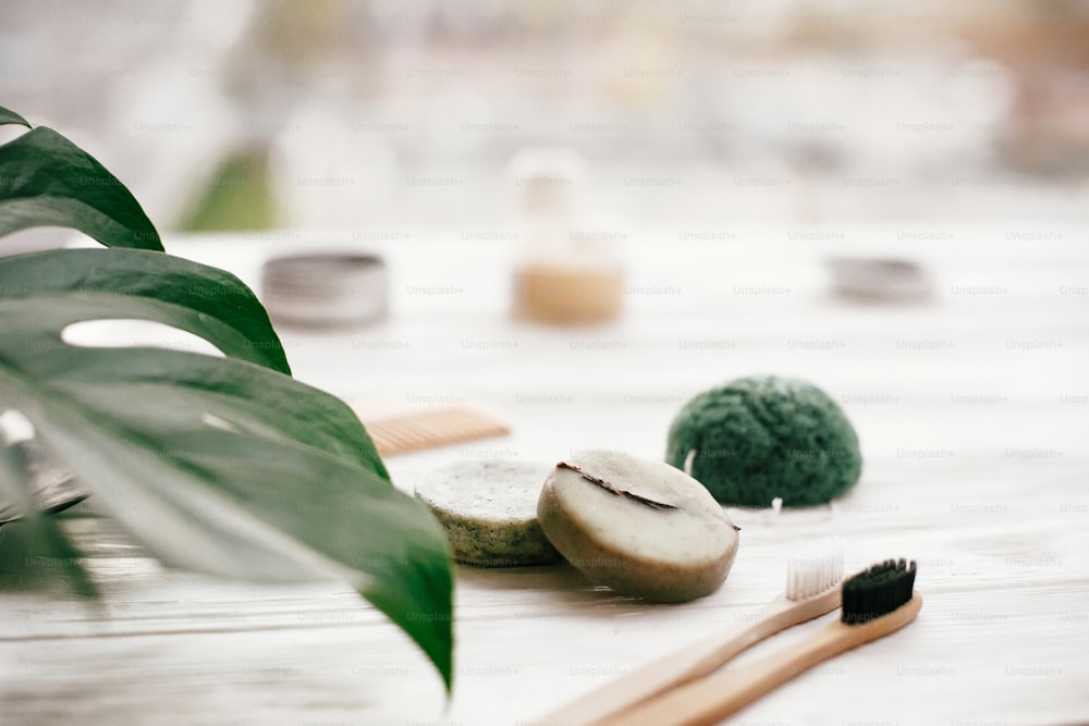 Eco friendly natural bamboo toothbrushes, shampoo bar, toothpaste in glass, wooden brush and konjaku sponge on white wood with green monstera leaves. Zero waste concept, plastic free