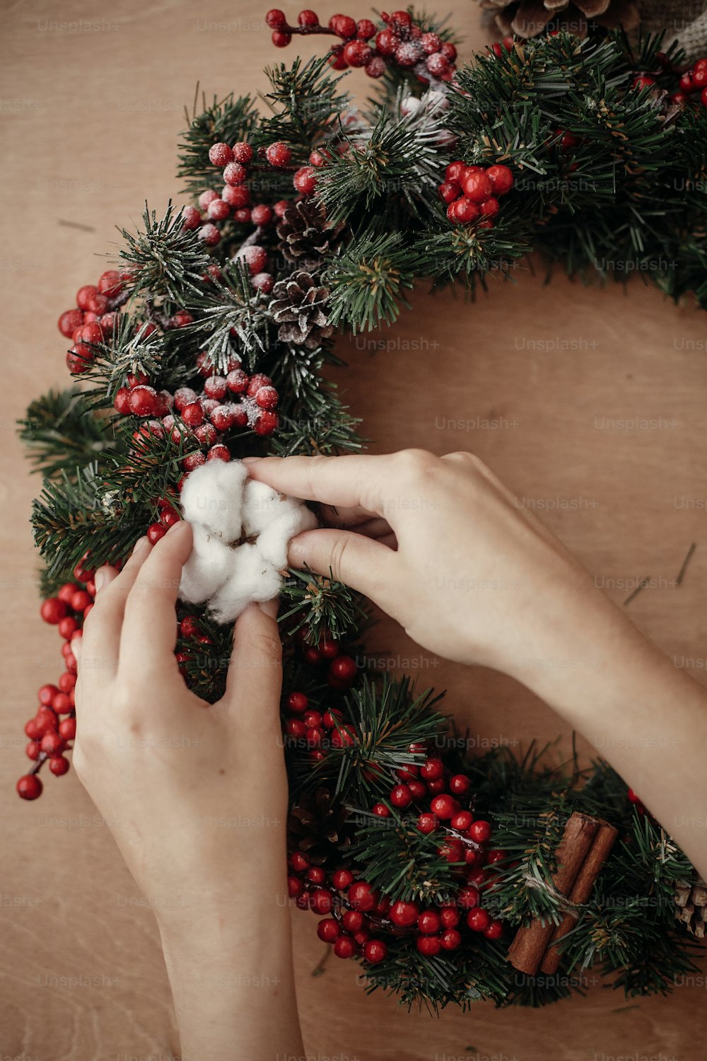 Hands making rustic christmas wreath, holding cotton at fir branches, red berries , pine cones on rustic wooden background. Atmospheric moody image at winter holiday workshop