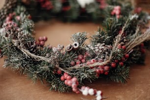 Details of rustic Christmas wreath, cinnamon and berries closeup. Fir branches with red berries,pine cones,ribbon, cotton on rustic wood. Atmospheric moody image at holiday workshop