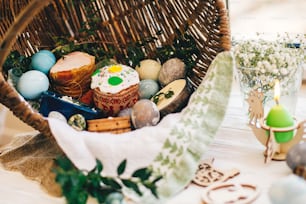 Traditional Easter basket with stylish eggs, easter bread, ham, sausage, butte, and flowers, candle on rustic wooden background. Happy Easter concept. Traditional food in rustic basket