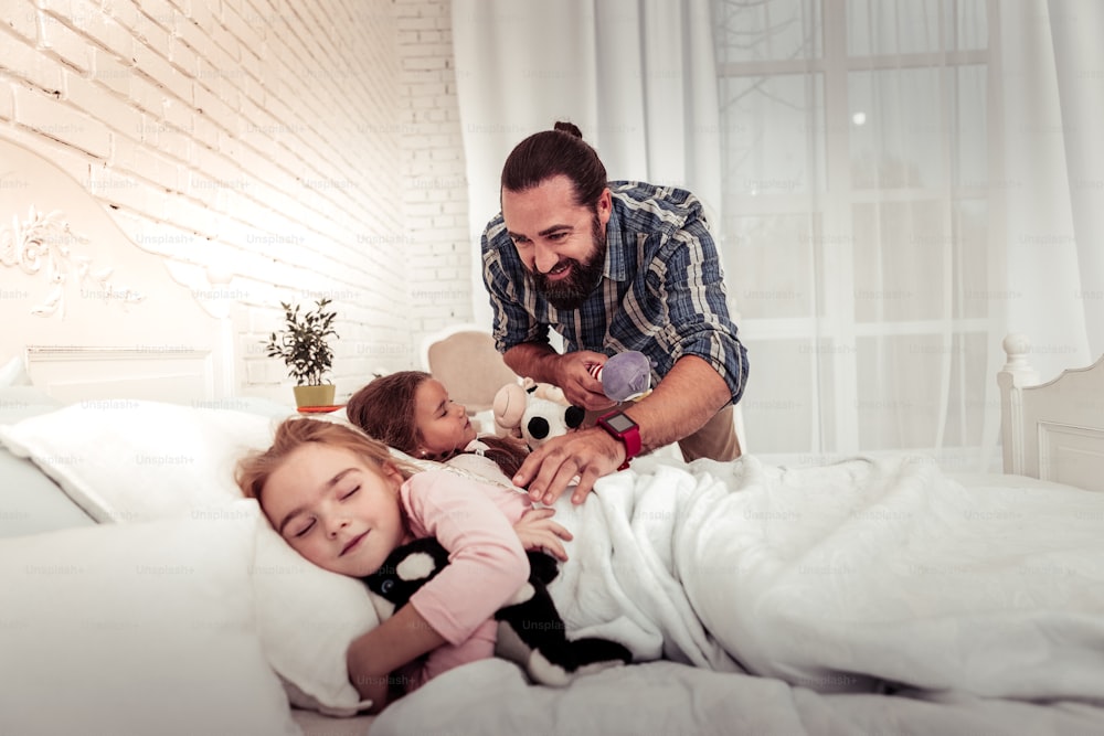 Wonderful father. Delighted bearded man standing at his daughters bed while caring about them