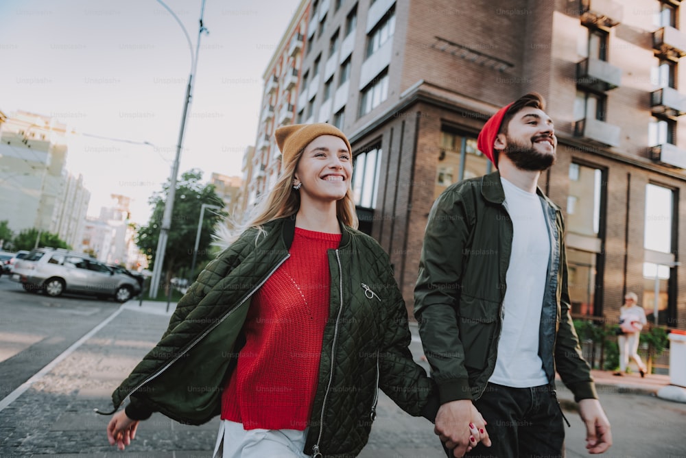 Waist up portrait of cheerful bearded man holding hand of his charming girlfriend. They looking away and smiling