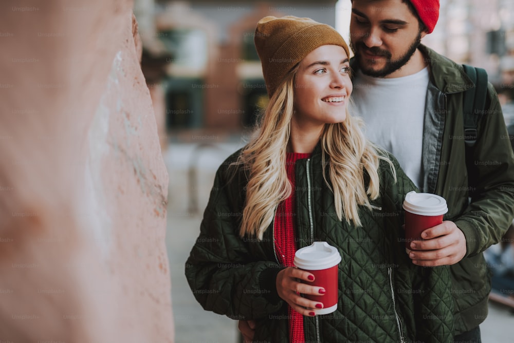 Waist up portrait of charming girl in hat holding cup of coffee while handsome bearded man looking at her with love and smiling