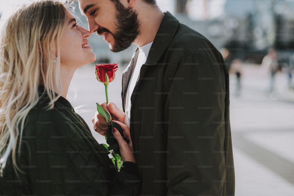 Retrato de vista lateral de un hombre guapo y barbudo que le da una rosa roja a su encantadora novia. Se miran con amor y sonríen