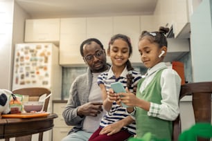 Modern daughters. Cute modern appealing daughters using their smartphone standing near father