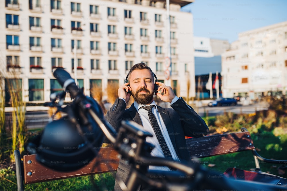 Businessman commuter with headphones and bicycle sitting on bench in city, listening to music.