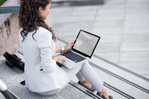 Young businesswoman sitting on stairs and using laptop outdoors.