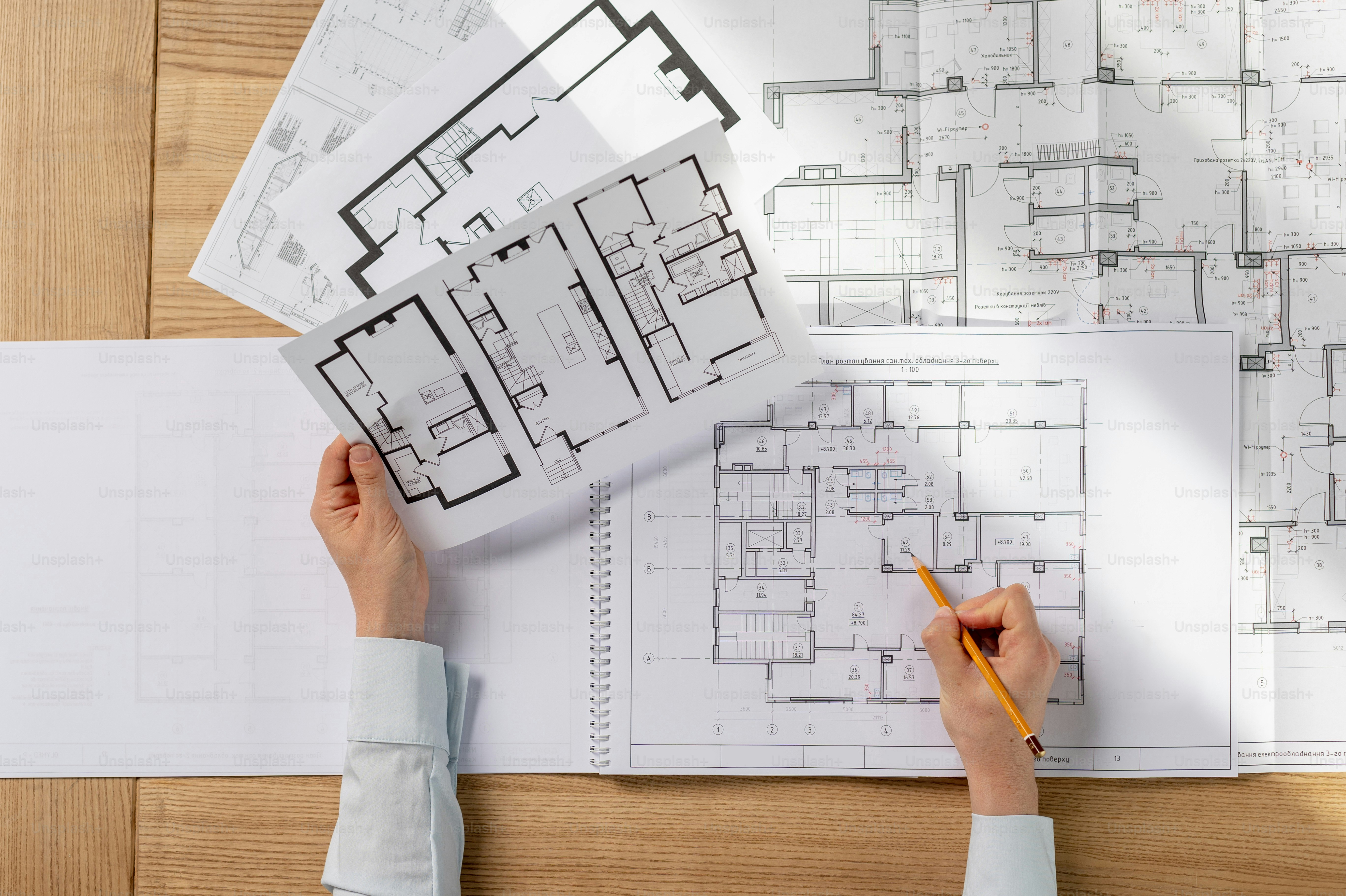 A person holding a pencil over architectural floor plans on a wooden desk
