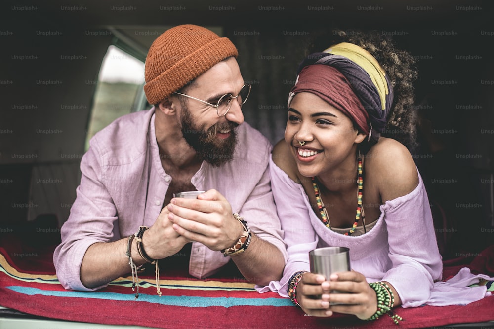 Concept of hippie lifestyle and rest. Waist up portrait of smiling ethnicity lady and male lover drinking tea in back trunk of retro van