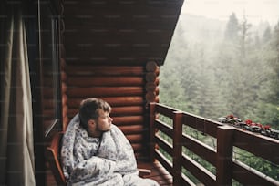 Homem viajante feliz descansando em cobertor na varanda de madeira com vista para bosques e montanhas. Espaço para texto. Hipster relaxando em cabana de madeira na floresta. Viagens e férias.
