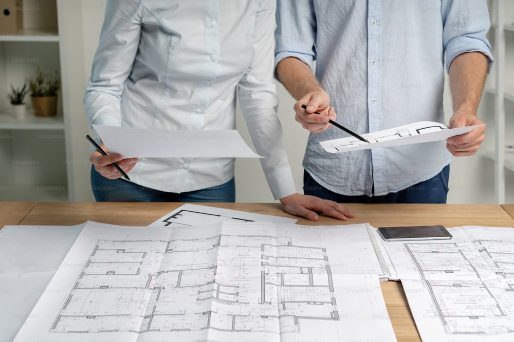 Cropped photo group of two person in formalwear shirt stand at the table in light loft interior workplace make modern renovation look at blank on room pointer on structure document by pencil