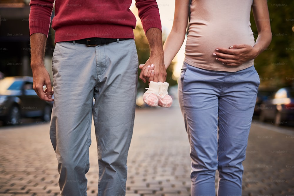 The happiest family. Close up of expectant parents posing with cute shoes for their child and sharing moment of closeness. Young pregnant woman gently touching her belly
