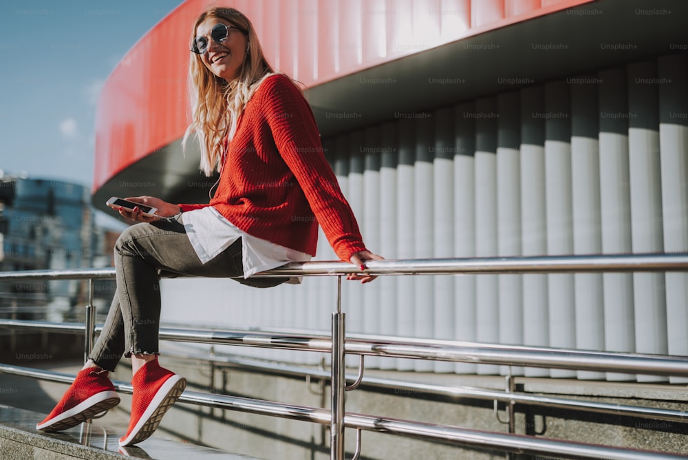 Full length portrait of beautiful girl in earphones holding smartphone while sitting on handrail. She is looking at camera and smiling