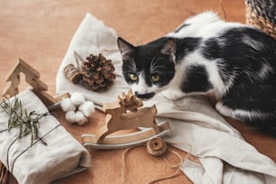 Süße Katze duftende rustikale Rentierspielzeug und stilvolles Weihnachtsgeschenk in Leinenstoff mit grünem Zweig auf Holztisch mit Tannenzapfen, Bindfaden, Baumwolle. Lustiger Moment. Einfache Öko-Geschenke