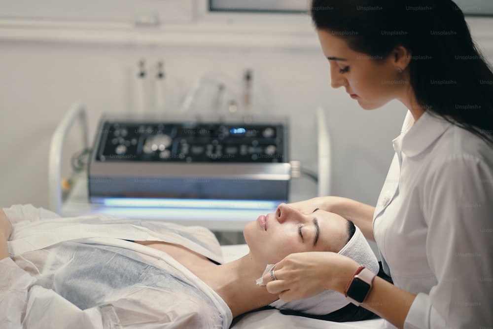 Mujer que miente durante el procedimiento cosmético mientras la hábil cosmetóloga hace el tratamiento facial
