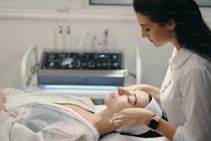 Woman lying during cosmetic procedure while skillful female cosmetologist making facial treatment