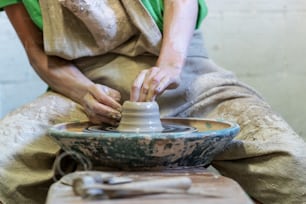 Foto recortada de cerca de la dama en su ropa de trabajo que se sienta dentro del producto de arcilla de cerámica de producción de espacio de trabajo por las manos disfruta del proceso