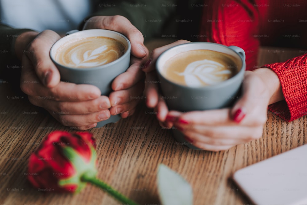 Close up of male and female hands holding cups of hot drink with foam. They sitting at the wooden table with red rose