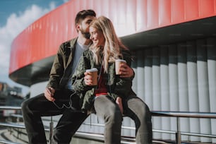 Portrait of bearded man kissing his charming girlfriend while she smiling. They holding cups of coffee and listening music with smartphone
