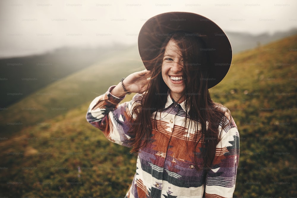 Portrait of happy young woman with backpack exploring mountains. Stylish hipster girl in hat smiling on top of mountains. Travel and wanderlust concept. Amazing atmospheric moment