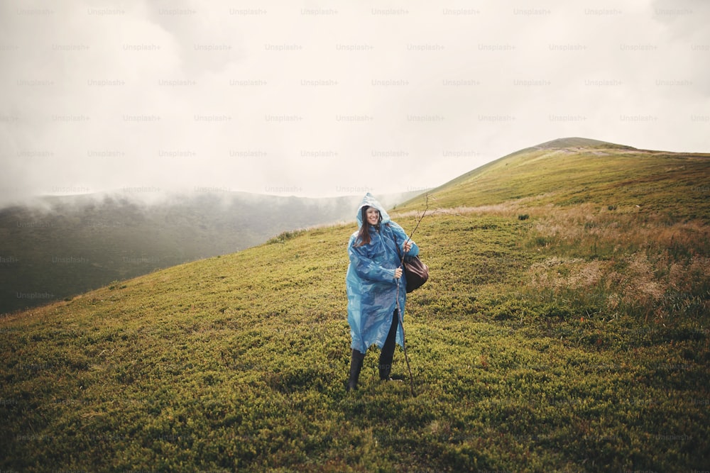 Elegante ragazza hipster in impermeabile blu e con zaino in piedi in cima a montagne nebbiose e nuvole. Giovane viaggiatrice divertente che sorride ed esplora le montagne. Viaggiare e voglia di viaggiare