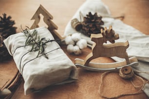 Stylish christmas rustic gift wrapped in linen fabric with green branch on wooden table with pine cones, reindeer, twine, cotton. Simple eco presents plastic free. Zero waste  holidays.