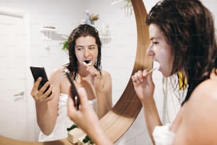 I social media influenzano. Giovane donna felice in asciugamano bianco che si lava i denti e guarda lo schermo del telefono in un bagno elegante allo specchio rotondo. Concetto di cura dei denti, routine quotidiana