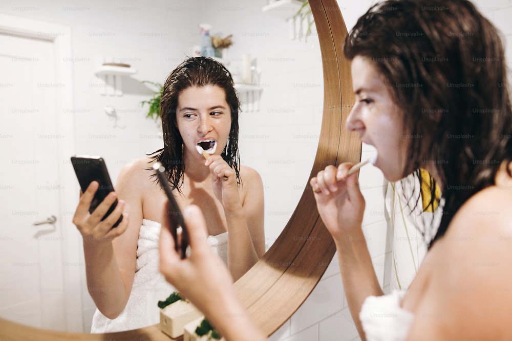 Social media affect. Young happy woman in white towel brushing teeth and looking at phone screen in stylish bathroom at round mirror. Tooth care concept, daily routine