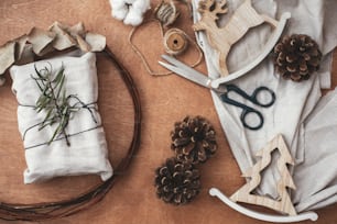 Stylish rustic gift wrapped in linen fabric with green branch on wooden table with pine cones, reindeer, scissors, twine, cotton. Flat lay. Simple eco presents plastic free. Zero waste christmas