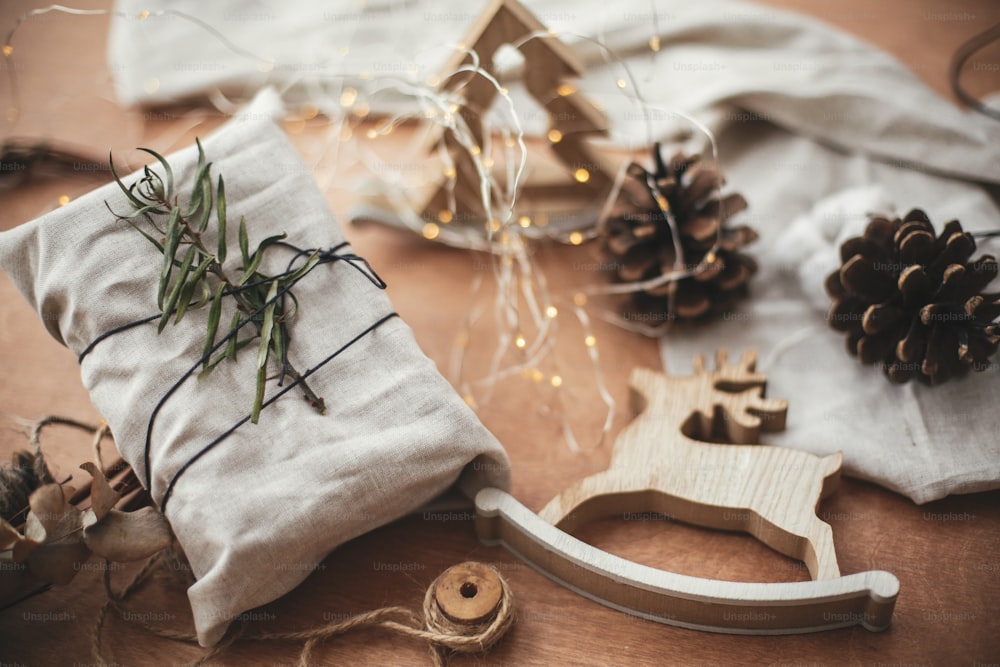 Feliz Navidad, concepto eco. Elegante regalo rústico envuelto en tela de lino con rama verde sobre mesa de madera con piñas, renos, luces. Regalos ecológicos sencillos. Vacaciones residuo cero.