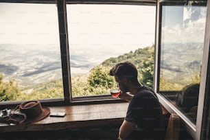 세련된 힙스터 여행자는 숲과 산의 아름다운 풍경을 배경으로 유리 컵에 뜨거운 차를 마시고 있다. 산의 나무 창문에서 바라보는 전망. 텍스트를 위한 공간입니다. 여행하다